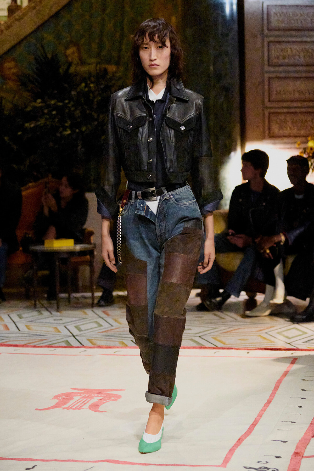 Model walking a runway in a black leather cropped jacket, patchwork denim jeans, and green flats at a fashion show venue.