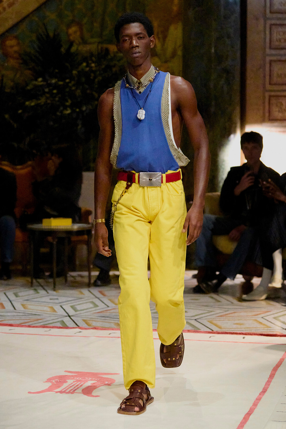 Male model on a runway wearing a blue sleeveless top over a checkered shirt, bright yellow pants, red belt, and brown sandals with a necklace and chain accessory.