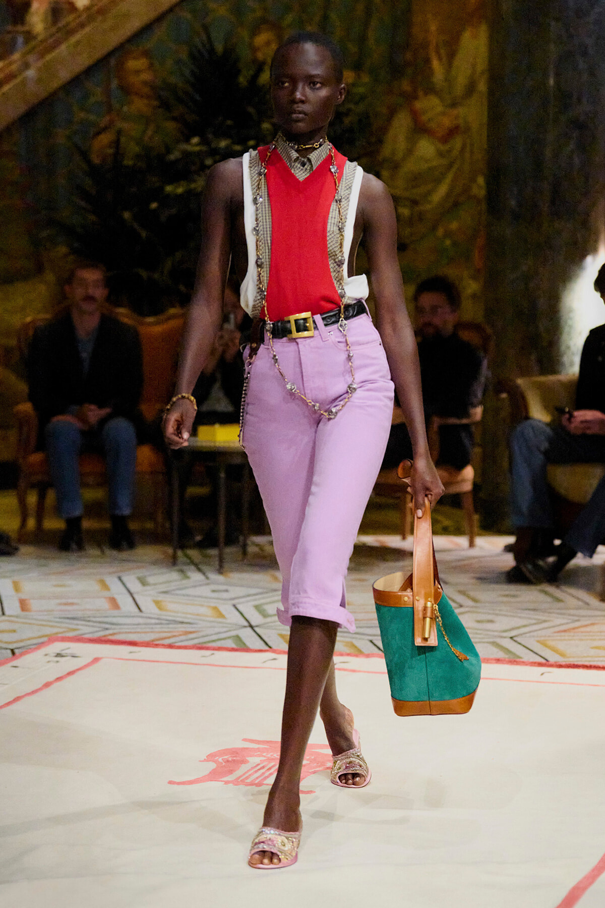 Model walking a runway in a red sleeveless top, lavender cropped pants, and gold chains; teal and tan tote bag in hand.