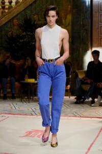 Model walks a runway in a white sleeveless halter top and royal blue pants with a black belt and two-tone heels.