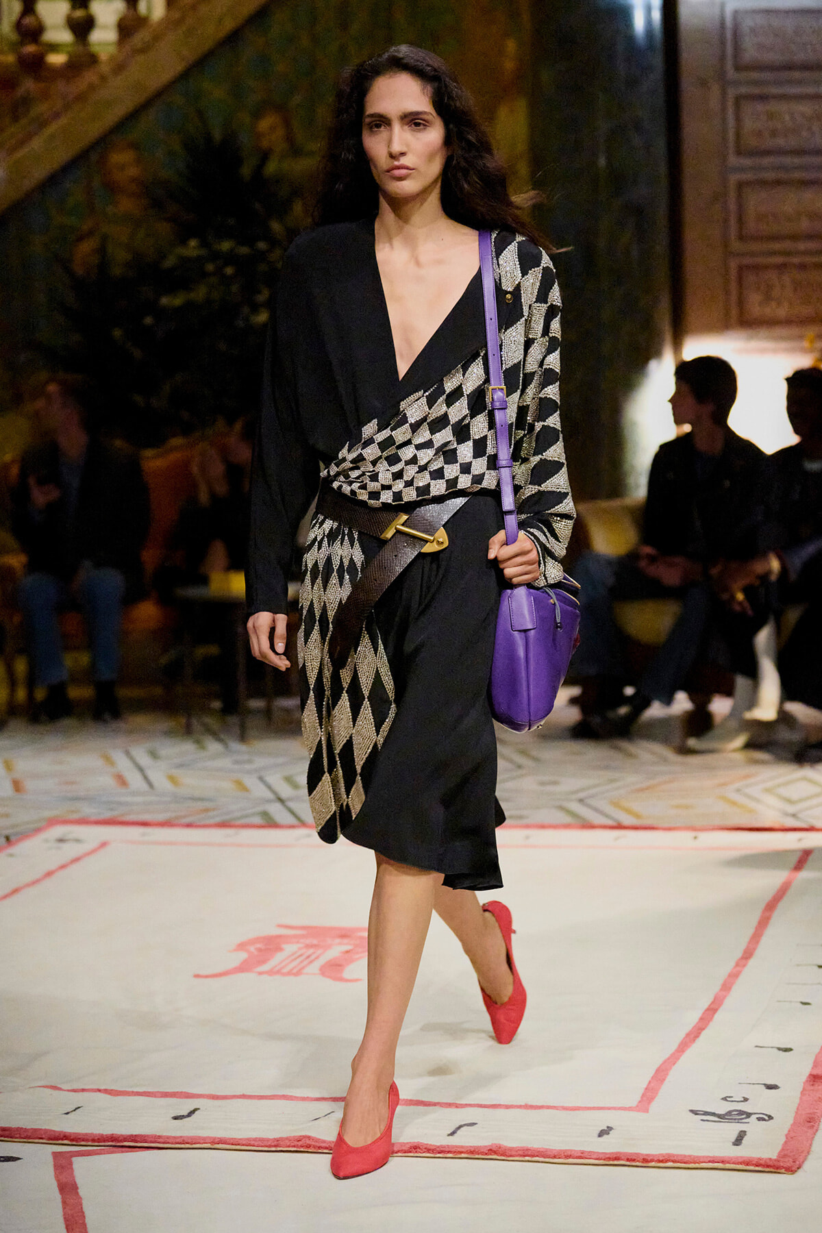 Model walking a runway in a black and white diamond-knit wrap, black dress, red heels, and a purple crossbody bag.