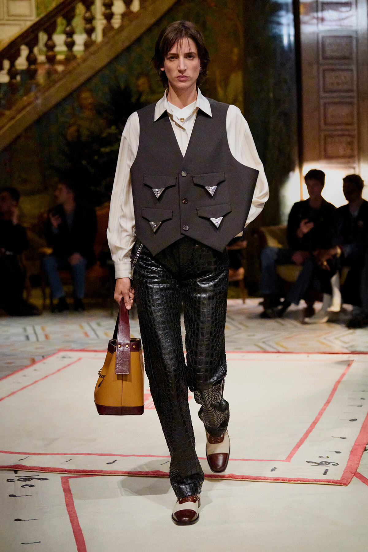 Model walking a fashion runway in a white shirt and dark vest with triangular pockets, paired with black textured pants and a tan handbag.
