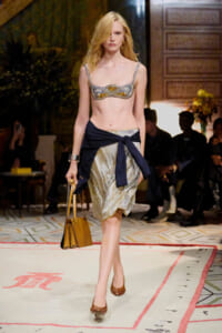Model walks the runway in a beaded metallic bra top and a shimmering wrap skirt with a navy sash, carrying a tan handbag.