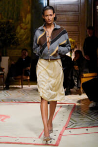 Model walking a runway in a color-block zip-front jacket and cream shimmering midi skirt, accessorized with layered necklaces, during a fashion show.