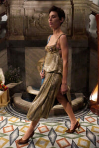 A woman in a shimmering gold evening gown with a beaded bodice walks across a colorful tiled floor in a grand interior.