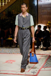 Male model walks a runway in a gray pinstripe suit with pastel mint sleeves and pink collar, accessorized with a long necklace and a blue-brown tote bag.