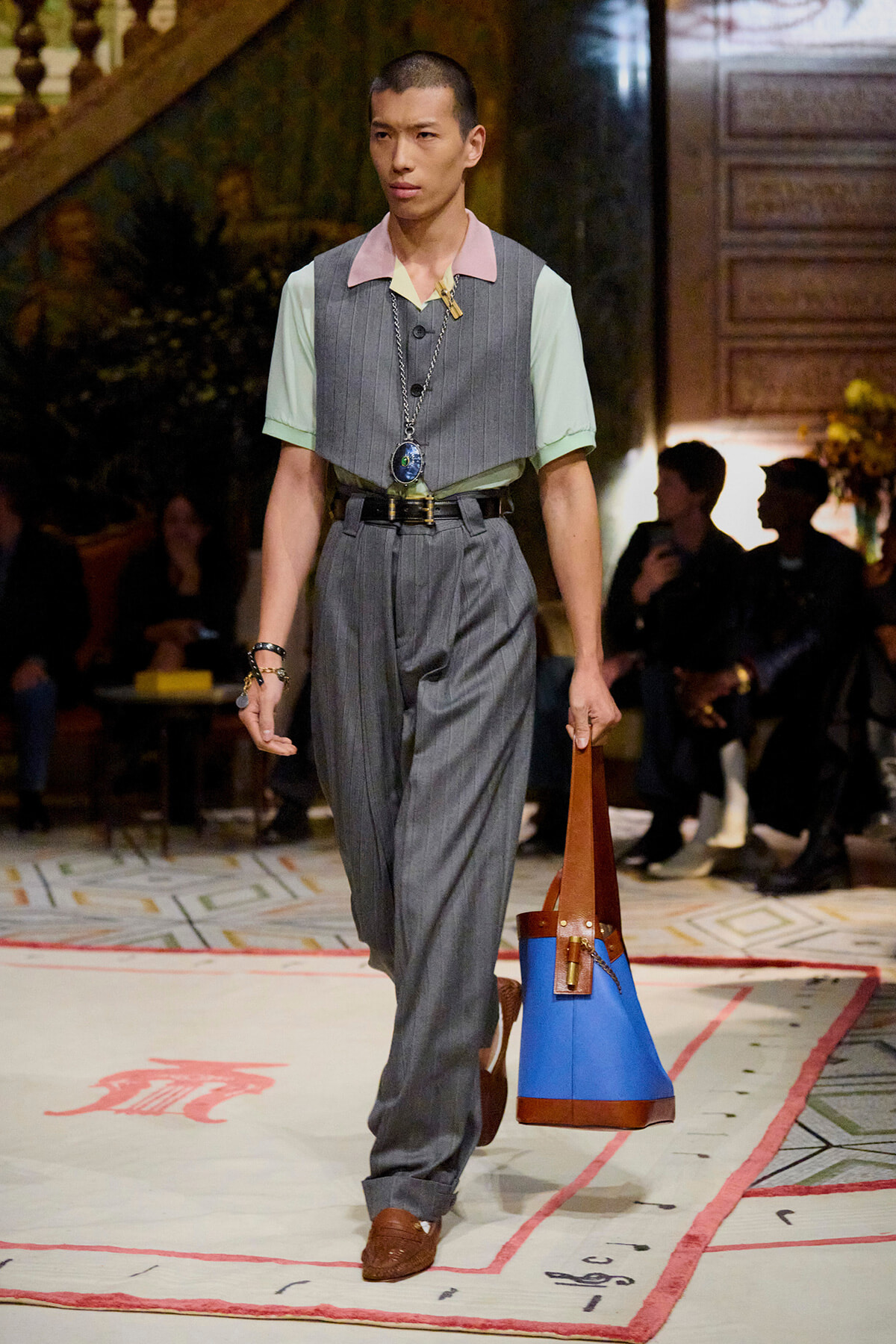 Male model walks a runway in a gray pinstripe suit with pastel mint sleeves and pink collar, accessorized with a long necklace and a blue-brown tote bag.
