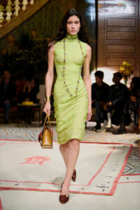 Model walking a runway in a lime-green ruched sleeveless dress, long beaded necklace, gold cuff, and orange patterned heels, carrying a tan handbag.