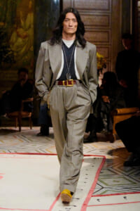 Male model with long black hair walks the runway in a gray pinstripe suit, black top, white tee, brown belt, and long necklace.