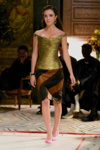 Model walking a runway in a gold off-the-shoulder top and color-blocked brown-black-tawny skirt, pink heels.