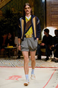 Male model walking a runway in a retro, color-blocked jacket over a yellow and brown V-neck sweater, tucked into gray pleated shorts with a bold gold belt and light blue socks plus white sneakers, on a patterned floor.
