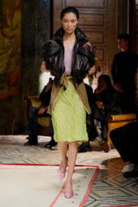 Model wearing an oversized black leather jacket over a lavender top and lime green pleated skirt struts down the fashion show runway, pink heels in view.