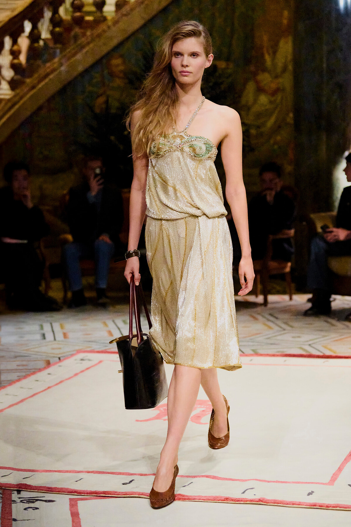 Model walking a fashion show in a gold halter dress, carrying a black tote and wearing brown heels.