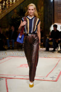 Model walks a runway in a sleeveless striped top, high-waisted brown crocodile-embossed pants, yellow pointed shoes, and carries a blue bag.