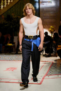 Male model walking a runway in a white sleeveless top, dark wide-leg pants, and a bold blue belt bag with a gold buckle.