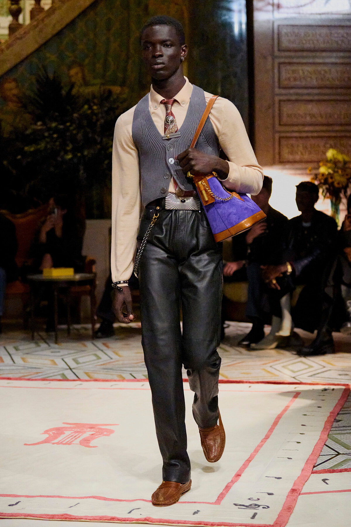 Male model walking a fashion show in a beige shirt, gray vest, black leather pants, brown loafers, carrying a purple handbag with tan straps.