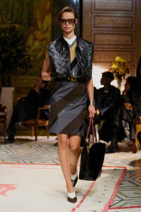 Runway look: model wears a black quilted leather vest over a color-block striped dress, white socks, brown shoes, and oversized sunglasses, carrying a black tote.
