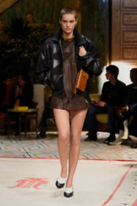 Model walking a fashion runway in a black oversized leather jacket over a brown mini dress, with white and black heels and a tan crossbody bag.