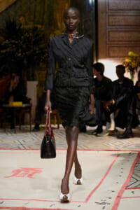 Model walks the runway in a structured black dress with puff sleeves, holding a black bag with red handles.