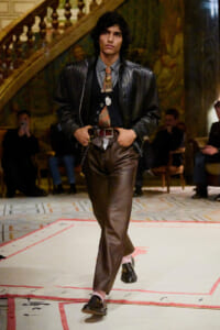 Male model walks a fashion runway in a black leather jacket, brown leather pants, and a patterned tie with vintage accessories.