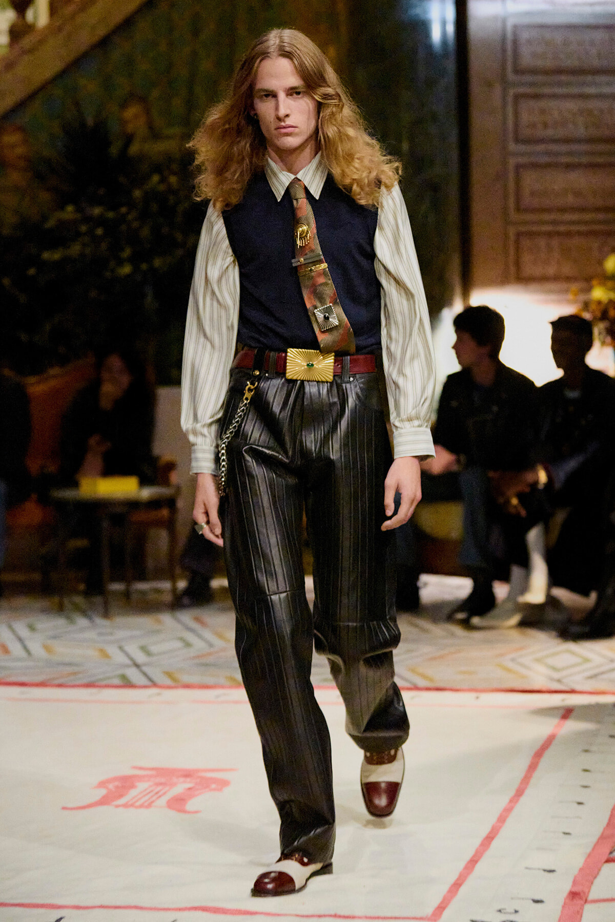 Male model walking a runway in a retro 1970s-inspired outfit: striped shirt with a navy vest, patterned tie, wide belt with buckle, pinstripe leather pants, and two-tone shoes.