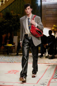 Male model walking a fashion runway in a gray blazer, striped shirt, pinstripe pants, and a red shoulder bag slung over his shoulder.