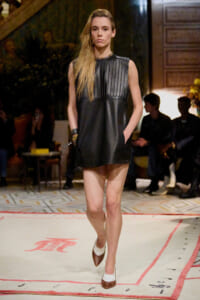 Model walking a runway in a sleeveless black leather dress with vertical pleats, hands in pockets, indoors at a fashion show