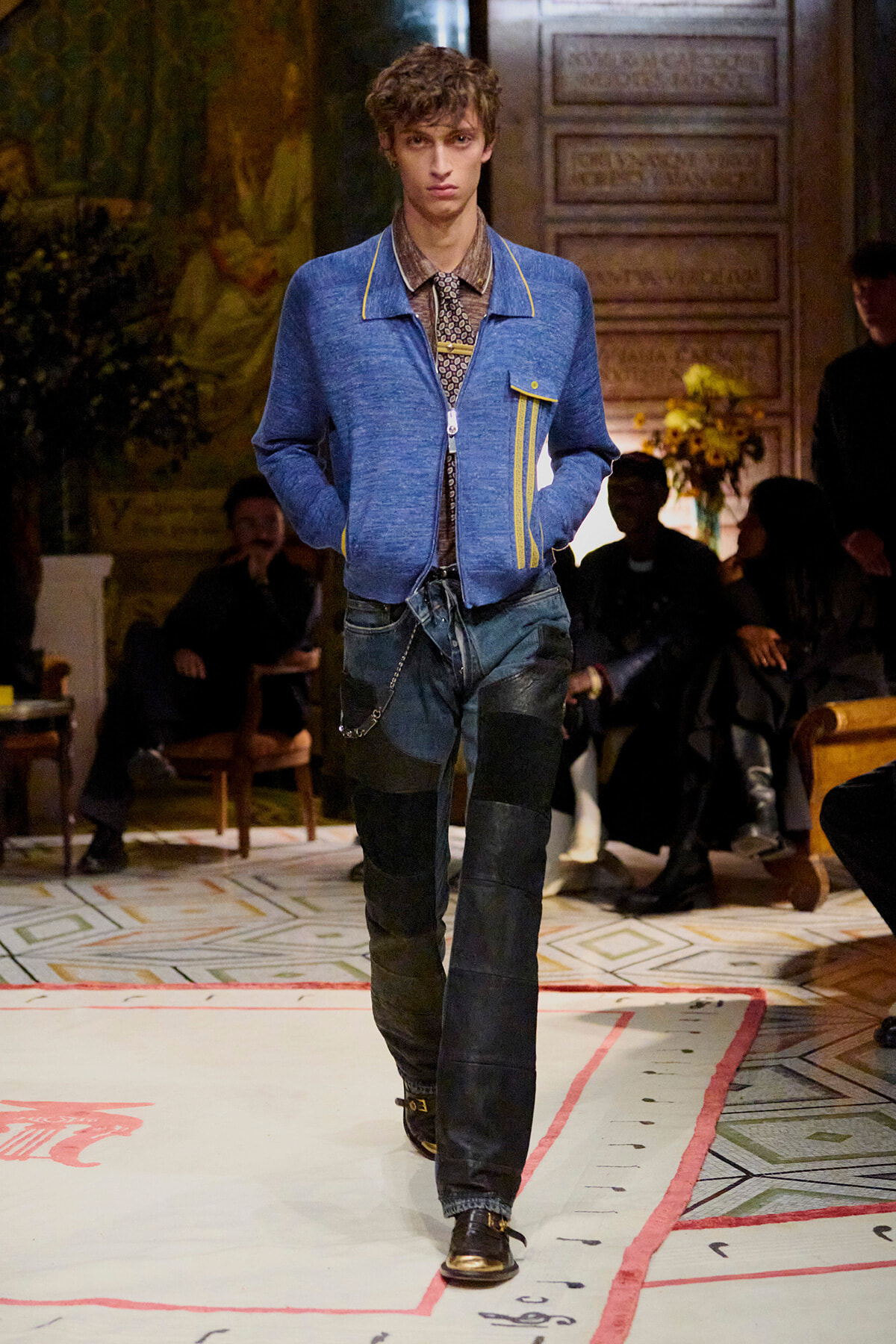 Male model walks a fashion show in a blue zip-front jacket with yellow piping, patterned shirt, and patchwork denim jeans on a runway.