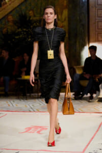 Model walks a fashion runway in a black ruched midi dress, accessorized with a large gold square pendant necklace and a brown leather tote.