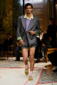 Male model walking a runway in a gray pinstripe blazer with green cuffs, lavender top, matching shorts, pink socks, and tan loafers carrying a crossbody bag.