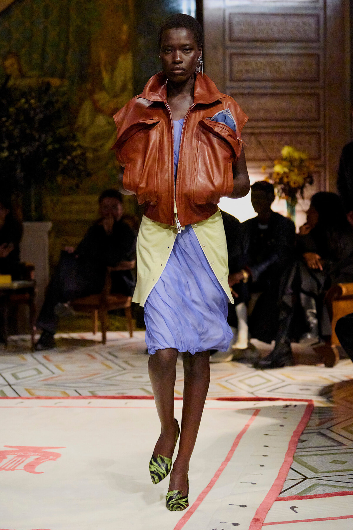 Model walking runway in an oversized brown leather jacket over a blue dress with a pale yellow layer and green zebra-print heels