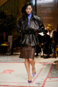 Model walking the runway in a black oversized leather jacket over a blue turtleneck and brown pencil skirt, paired with purple heels.