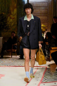 Runway model in a navy oversized blazer with a mint-green collar, black shorts, blue socks, and gold shoes, carrying a tan bag.
