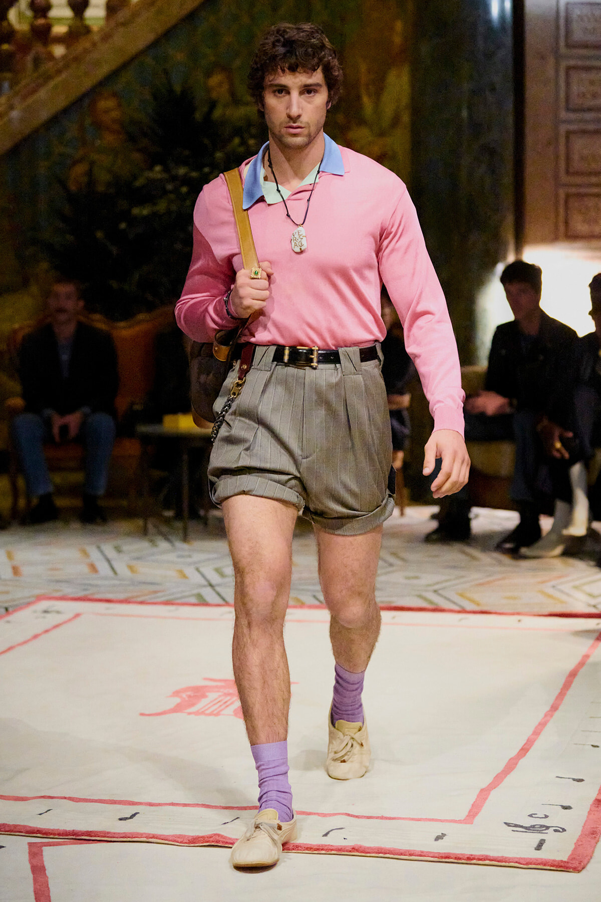 Male model walks a runway in a pink long-sleeve polo, gray pinstriped shorts, lavender socks, and beige shoes, carrying a shoulder bag and necklace.