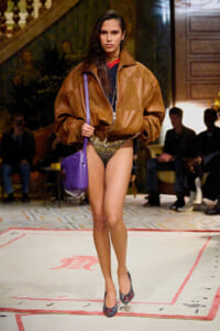 Model walking the runway in an oversized brown leather jacket, glittery bikini bottoms, and a purple shoulder bag.