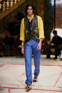 Male model on a runway wearing a bright yellow shirt with an ornate black embroidered vest and blue trousers.