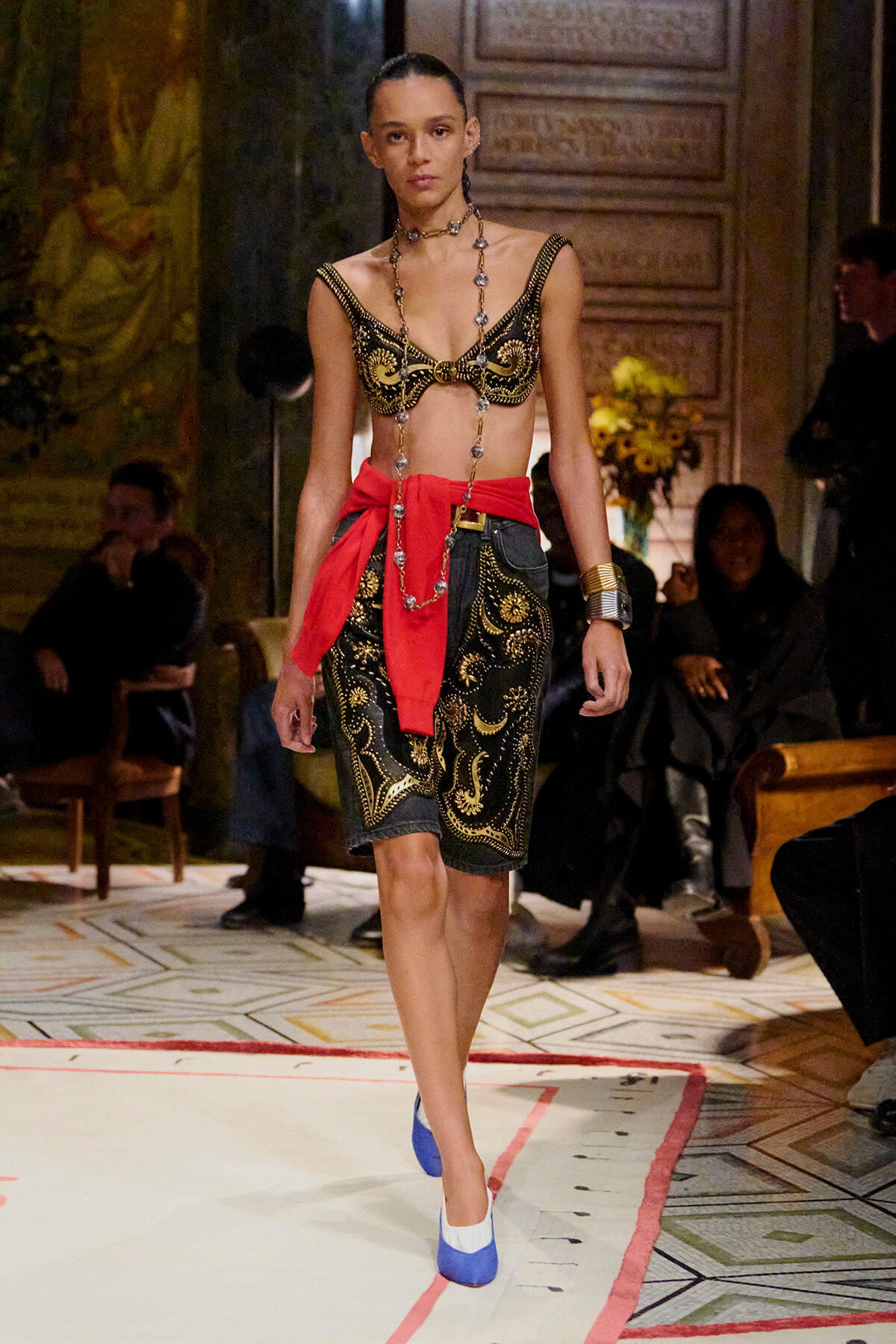 Runway model in black and gold embroidered bralette with matching paisley shorts, red waist sash, long beaded necklace, and blue-and-white heels.