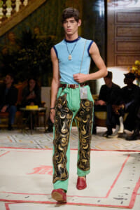 Model walking a fashion runway in a light blue sleeveless top and bright green pants with black-and-gold embroidery, red shoes, and a pendant necklace with a belt.