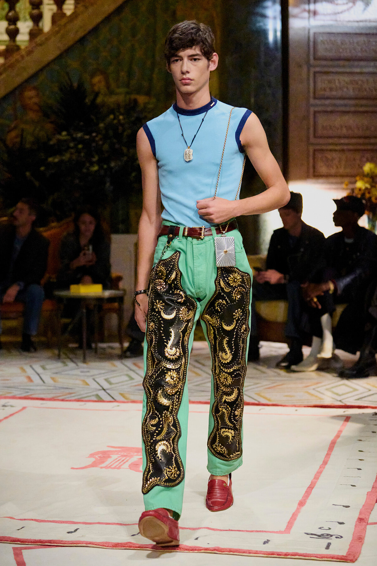Model walking a fashion runway in a light blue sleeveless top and bright green pants with black-and-gold embroidery, red shoes, and a pendant necklace with a belt.