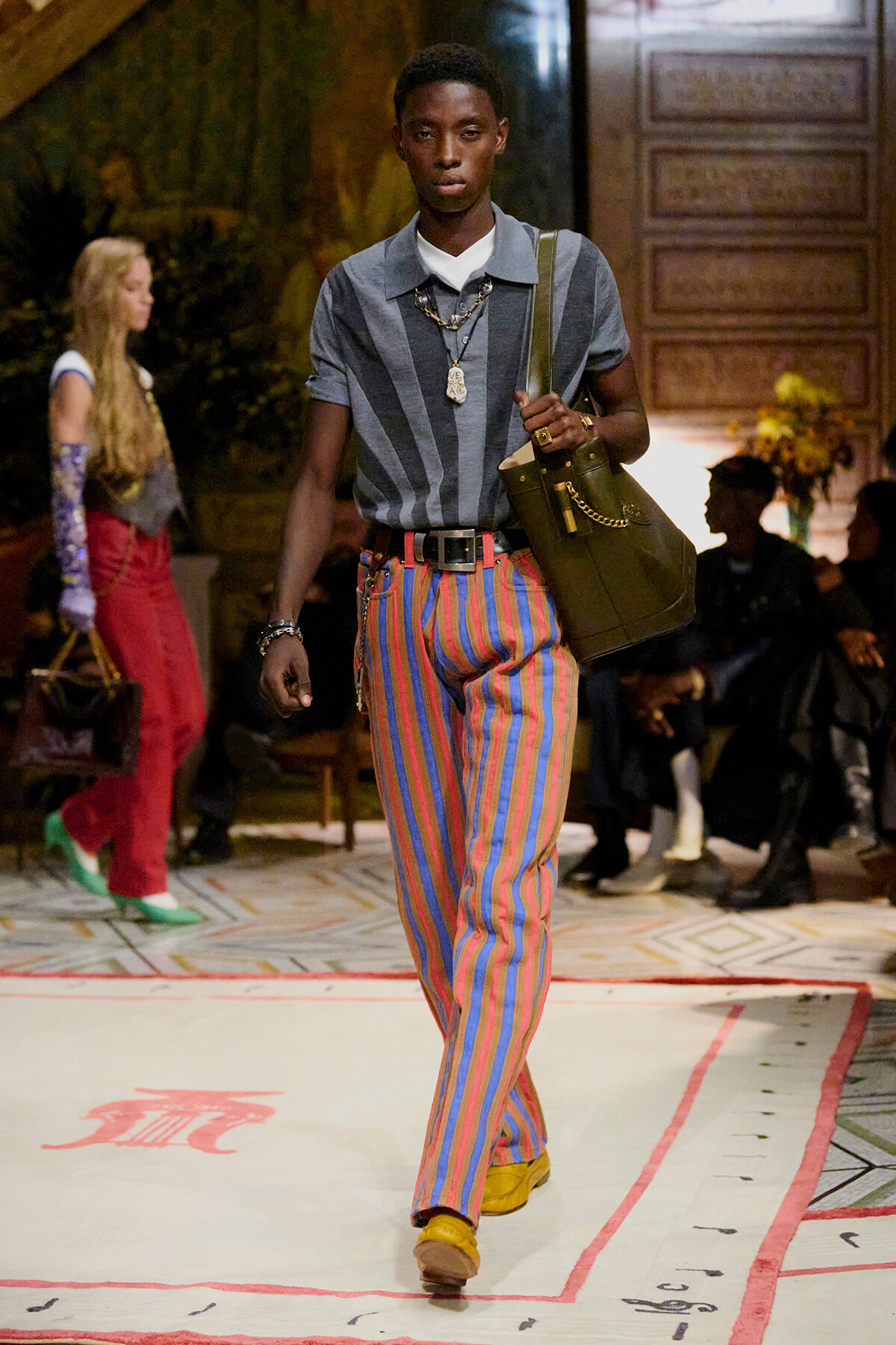 Male model walks a fashion runway wearing a blue-gray striped polo, multicolored striped pants, yellow shoes, and a green messenger bag. jewelry includes layered necklaces and bracelets.