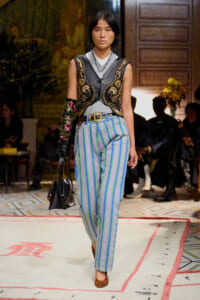 Model on a runway wearing a black ornate embroidered vest over a gray top and blue-green striped trousers, carrying a black handbag.