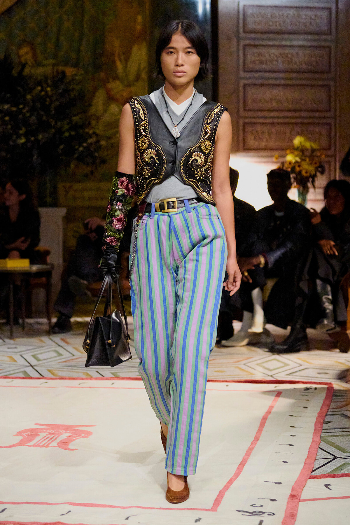 Model on a runway wearing a black ornate embroidered vest over a gray top and blue-green striped trousers, carrying a black handbag.