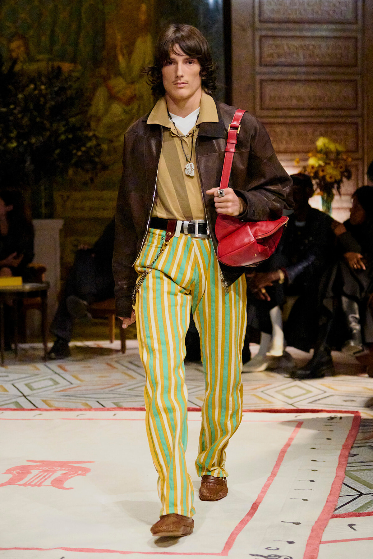 Male model on a runway wearing bold yellow and green striped pants, a brown jacket, beige shirt, and a red crossbody bag while walking.