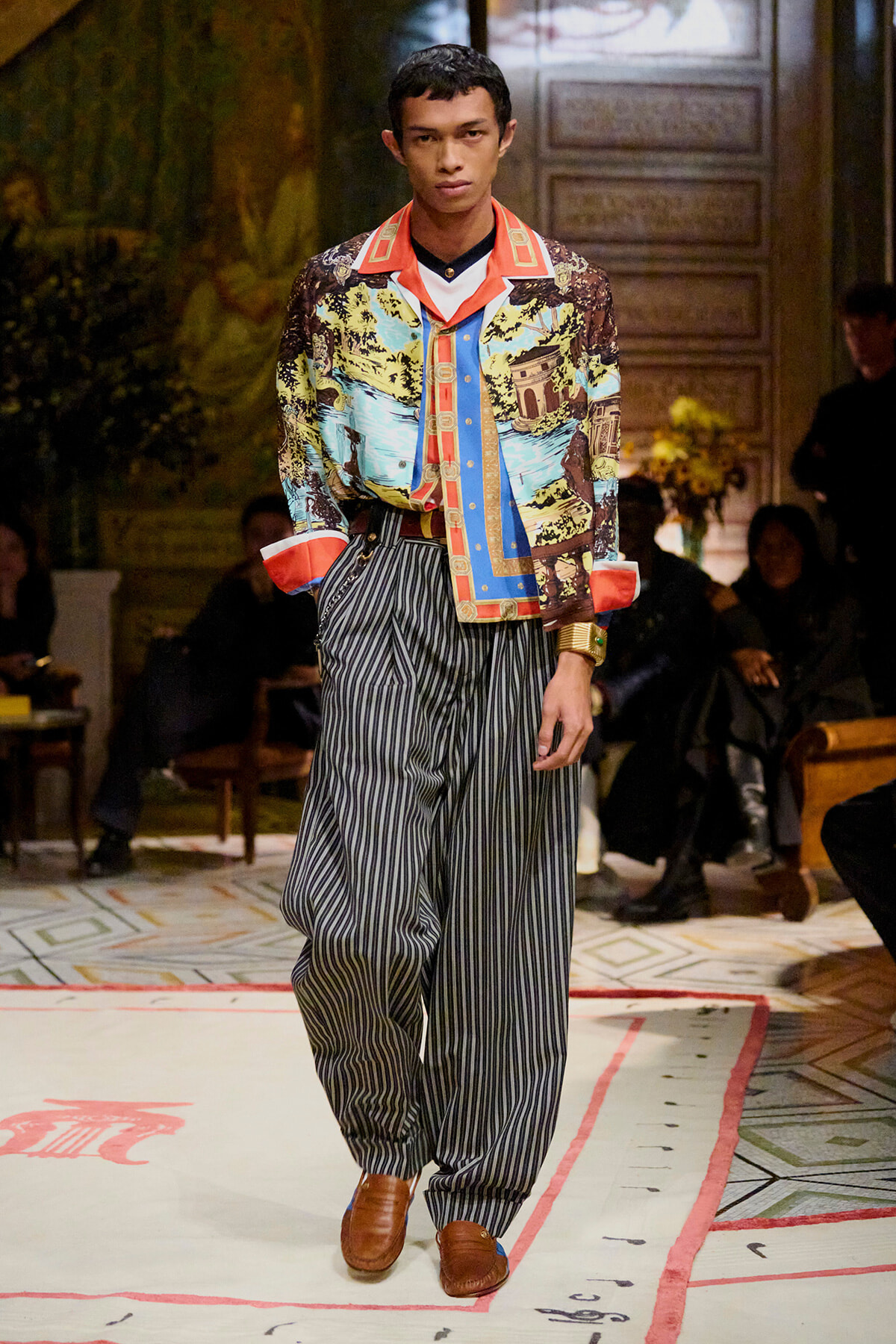 Male model on a runway wearing a vibrant printed shirt and baggy striped trousers with brown shoes.