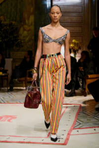 Model on a runway wearing a metallic silver beaded bralette and colorful vertical-striped pants, carrying a burgundy handbag.