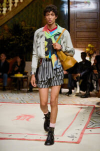 Male model walks a runway in a colorful layered outfit with striped gray shorts, black boots, pink socks, and a tan leather shoulder bag.
