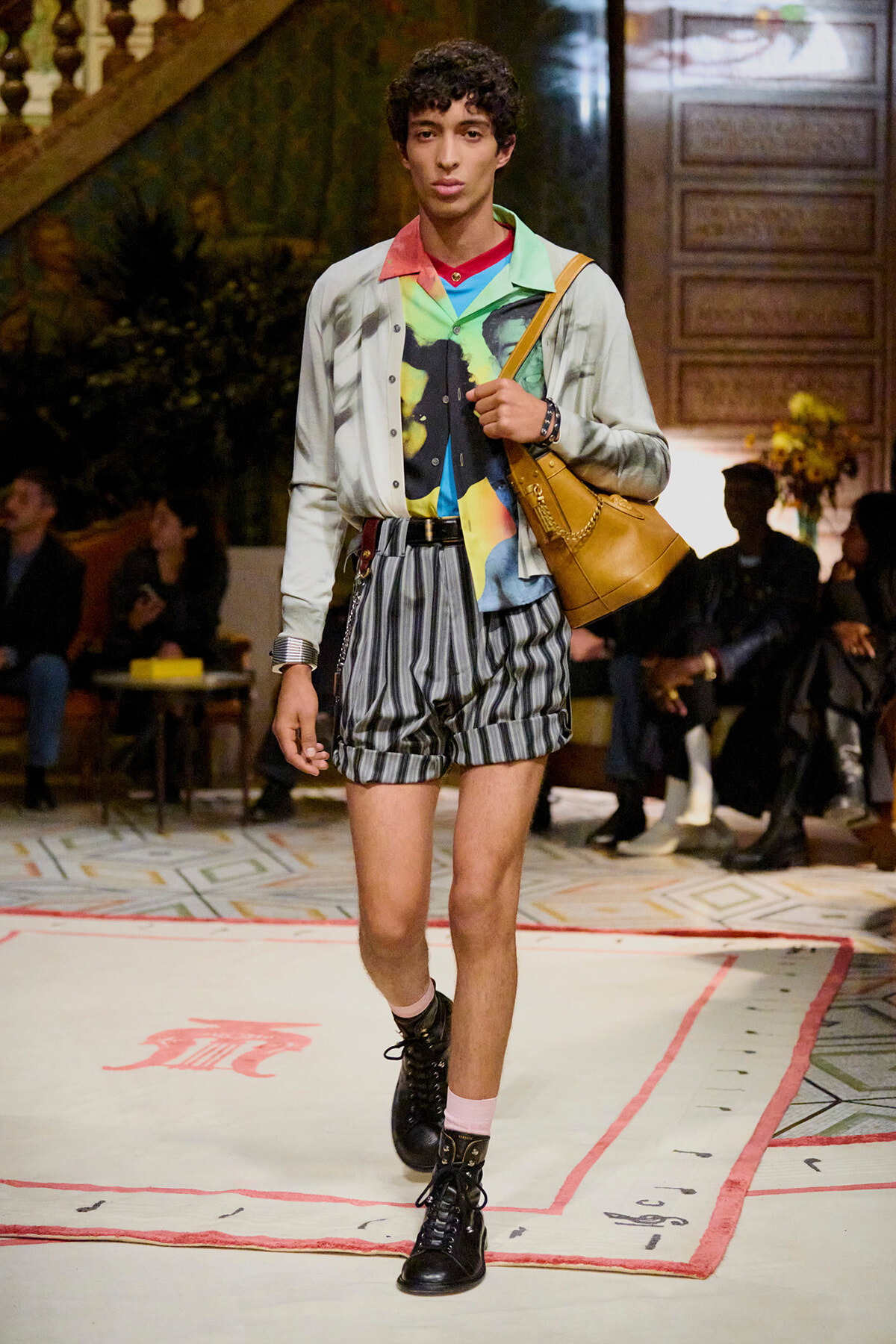 Male model walks a runway in a colorful layered outfit with striped gray shorts, black boots, pink socks, and a tan leather shoulder bag.