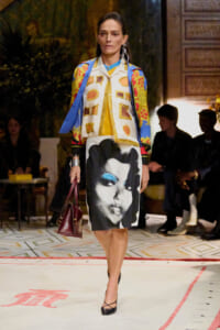 Model walking a runway in a colorful patterned jacket and a knee-length skirt featuring a large black-and-white portrait.