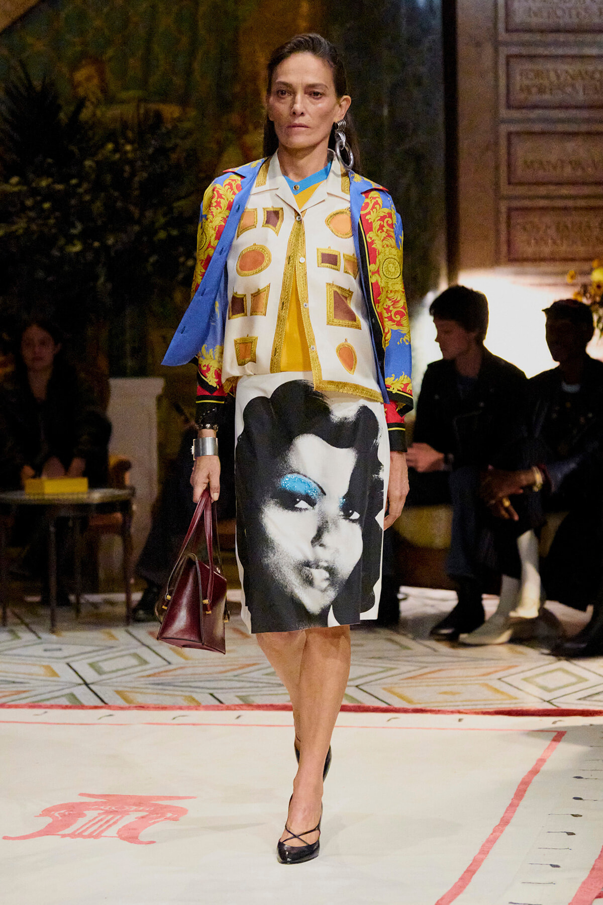 Model walking a runway in a colorful patterned jacket and a knee-length skirt featuring a large black-and-white portrait.