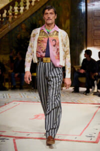 Male model walks a fashion runway in a retro outfit: floral cardigan over a tropical print shirt, striped trousers, and a wide gold belt.